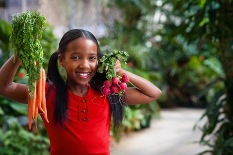 A smiling girl with pigtails in a red shirt holds up some carrots and radishes