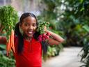A smiling girl with pigtails in a red shirt holds up some carrots and radishes