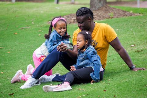 A father and his two children look at a phone screen sitting outside on a grassy hill.