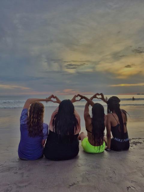Four youth sit on the beach next to each other. Their arms are linked and raised above their heads, with their hands forming hearts.