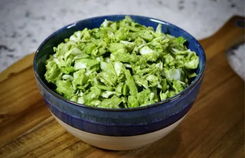 A blue-and-white bowl filled with finely chopped green broccoli florets and stems, lightly coated in a creamy green dressing, sitting on a wooden cutting board.