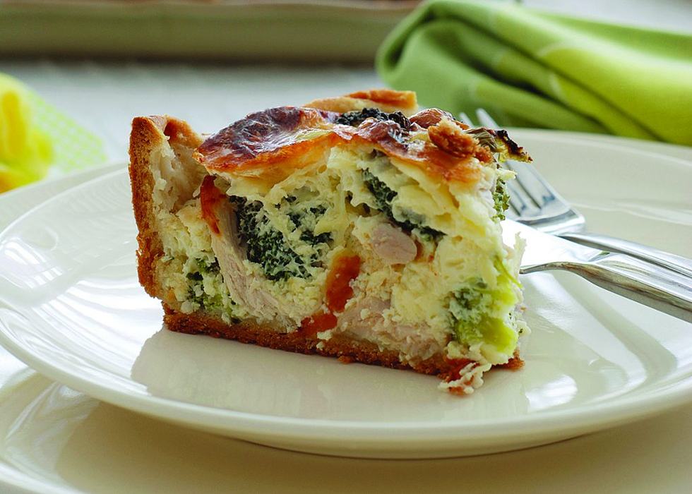 A slice of chicken and broccoli quiche with a golden crust on a white plate with a fork.