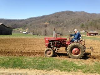 Person driving red tractor plowing a field, barns and wooded hills in background