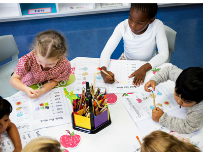 Five children seated at a table completing worksheets and coloring with a center pencil caddy