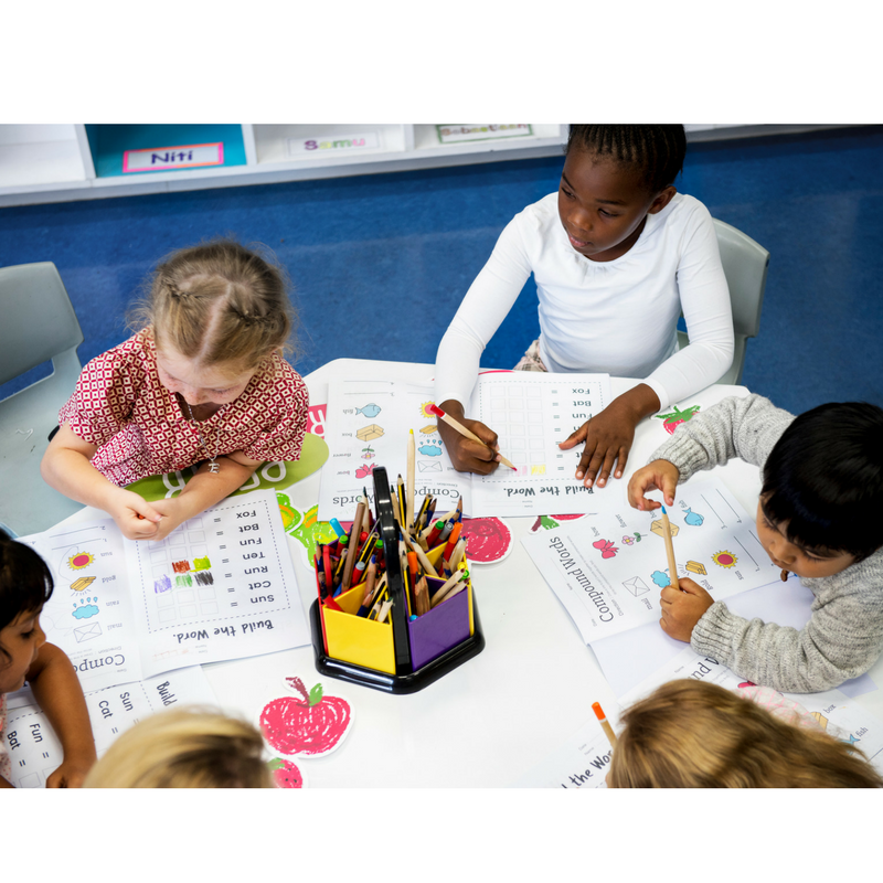 Five children seated at a table completing worksheets and coloring with a center pencil caddy