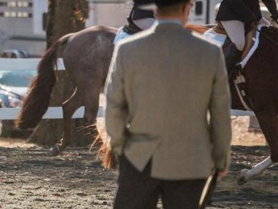 Person in jacket and hat holding clipboard watching riders on horses in arena