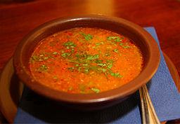 Tomato soup with green herbs in a brown bowl sitting on a blue cloth on a dining table. 