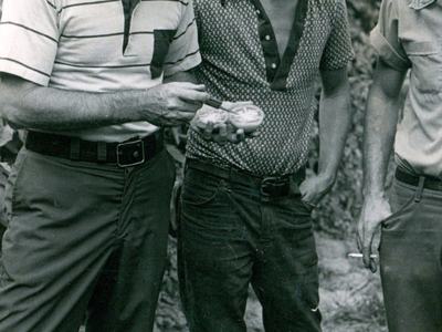Three men standing; left man shows sliced fruit to middle man wearing "CAT Diesel Power" cap.
