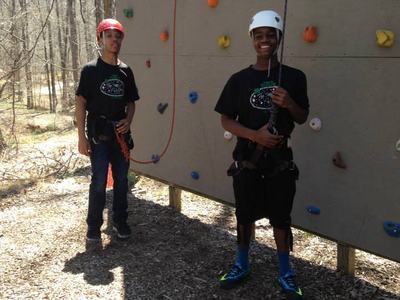 Two youths in helmets and harnesses standing by an outdoor rock-climbing wall.