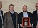 Four men standing side by side; second from right holds a framed award plaque.