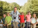 Adult with binoculars and five children on a wooden railing by a pond