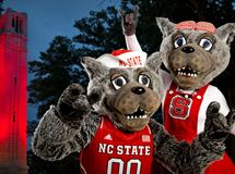 Two NC State wolf mascots in red uniforms at night, both flashing the wolf hand sign