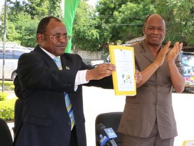 Two men at an outdoor event; man on left holds up a framed document while man on right claps.