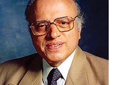 Older man wearing glasses, beige suit and patterned tie against a blue studio background