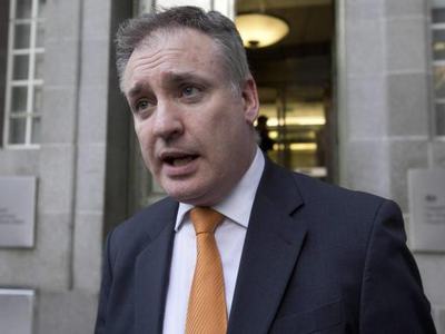 Man in dark suit and orange tie speaking outside a stone, institutional-style building