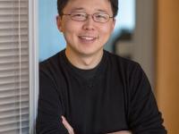 Person with glasses in black sweater standing with arms crossed by a glass office partition
