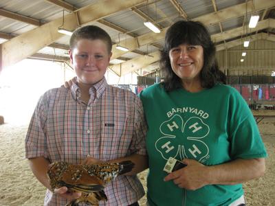 Boy holding patterned chicken beside woman in green "BARNYARD" 4-H shirt inside barn