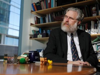 Bearded man in suit and tie seated at desk, looking to the side with bookshelves behind
