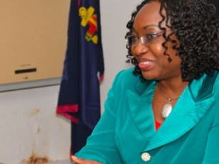 Woman in teal jacket seated at a desk, speaking and gesturing, flag in background