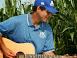 Man in blue cap and polo playing acoustic guitar while seated in a cornfield.