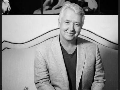 Middle-aged man in blazer seated on a sofa with hands clasped, flowers on a table nearby.