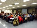 Large audience seated at tables listening to a presenter with a projected slide in a community hall