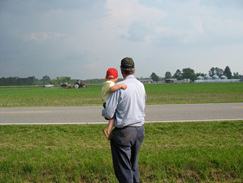 Man carrying child standing by roadside facing open farm field with equipment in distance