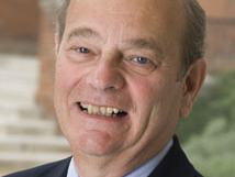 Older man in suit and green tie smiling, steps and brick building behind him