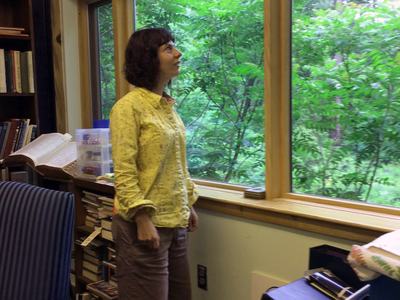 Person standing by large window in a book-filled room, looking outside at trees