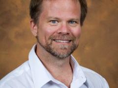 Man with short brown hair and beard wearing a white collared shirt against a mottled brown background
