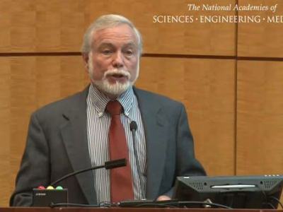 Man in suit speaking at podium; text "The National Academies of SCIENCES · ENGINEERING · MEDICINE" visible