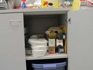 Open lower cabinet under counter holding stacked food containers, cartons, and a blue storage bin