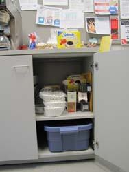 Open lower cabinet under counter holding stacked food containers, cartons, and a blue storage bin