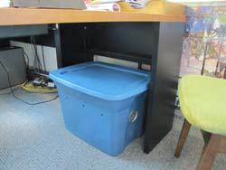 Blue plastic storage bin with lid under office desk beside green chair