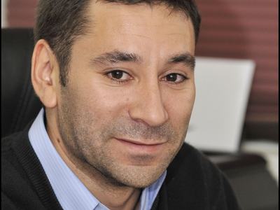 Man with short dark hair wearing blue striped shirt and dark sweater, seated indoors