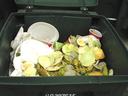 Green compost bin filled with fruit peels, cut melons, and disposable plates