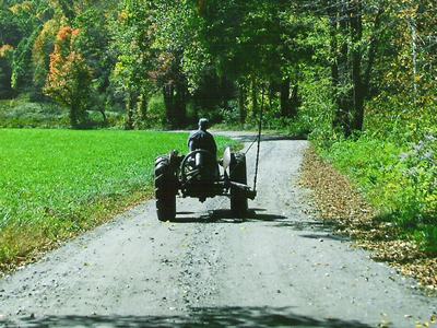 Person driving a tractor down a tree-lined dirt road beside green fields