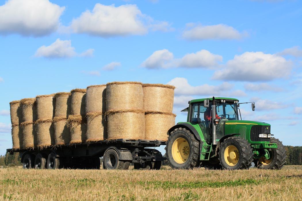 Green tractor pulling trailer loaded with round hay bales across harvested field