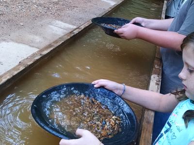 Children panning gravel and small rocks in black pans at a long water sluice