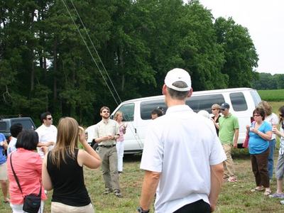 Group of people standing in a field listening to a guide near white vans