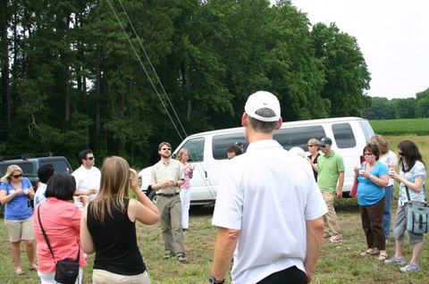Group of people standing in a field listening to a guide near white vans