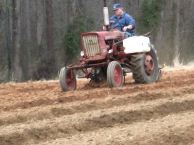 Person driving red tractor plowing furrowed field with trees in background