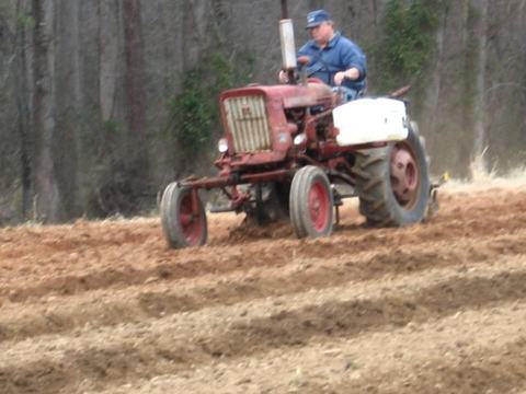 Person driving red tractor plowing furrowed field with trees in background