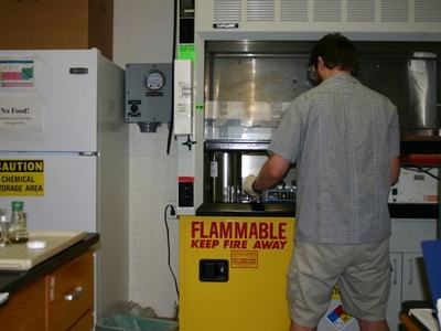 Person working at a lab fume hood above a yellow cabinet labeled "FLAMMABLE KEEP FIRE AWAY"