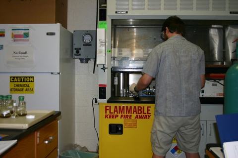 Person working at a lab fume hood above a yellow cabinet labeled "FLAMMABLE KEEP FIRE AWAY"
