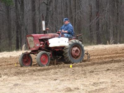 Man driving red tractor plowing a field with leafless trees in background.
