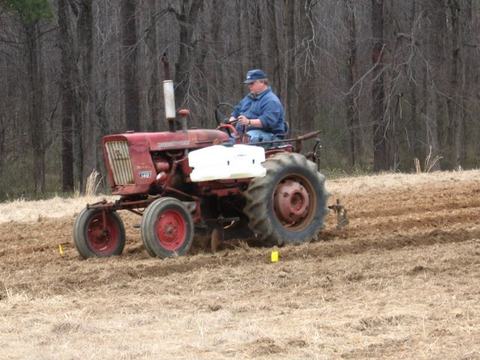 Man driving red tractor plowing a field with leafless trees in background.