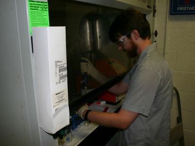 Man with goggles and gloves pipetting inside a lab hood; "FIRSTAID" sign.