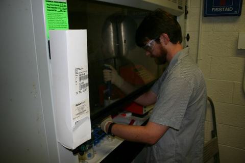 Man with goggles and gloves pipetting inside a lab hood; "FIRSTAID" sign.
