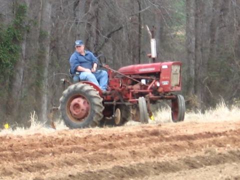 Man driving red tractor across plowed field with trees in background